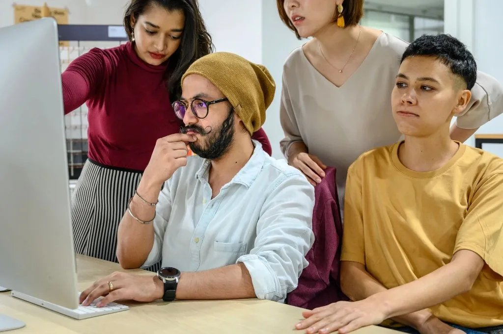 Four people gathered around a desktop computer, reviewing what is on the screen.